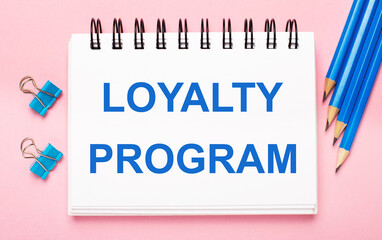 On a light pink background, light blue pencils, paper clips and a white notebook with the text LOYALTY PROGRAM