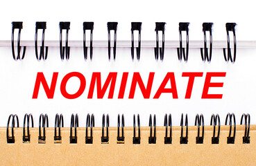 Text NOMINATE on white paper between white and brown spiral notepads.