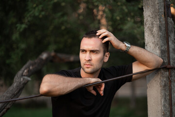Portrait of a young handsome man with unshaven bristles in a black T-shirt leaning on a wire