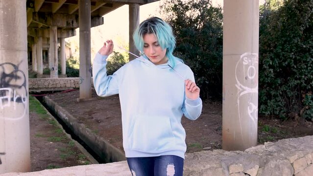 Smiling teenager wearing hoodie and staying against bridge pillars. Blue haired teen girl stays outdoors against bridge pillars. Clothing mockup