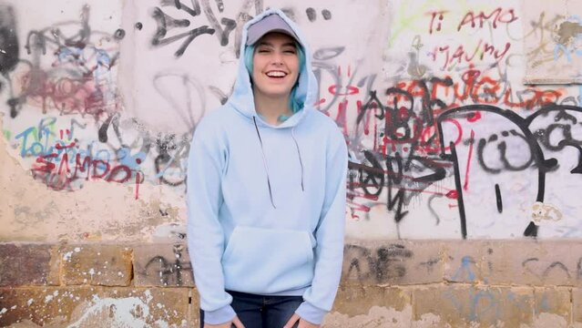 Laughing Teenager in light blue oversize hoodie and jeans. Blue haired teen girl stays outdoor against graffiti wall. Clothing mockup