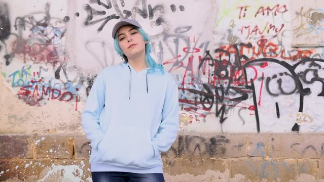 Teenager in light blue oversize hoodie and jeans taking off hood. Blue haired teen girl stays outdoor against graffiti wall. Clothing mockup