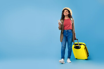 Young black woman with suitcase holding smartphone, booking hotel or vacation online, using web...