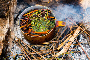Survival in wild nature. Campfire background. Pan with cherry tea sticks over fire. Cooking tea...