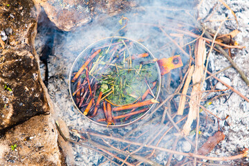 Survival in wild nature. Campfire background. Pan with cherry tea sticks over fire. Cooking tea drink at fire in wild.