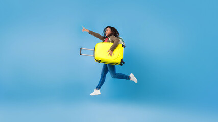 Happy young black woman jumping with suitcase and pointing at blank space for travel agency ad on blue background