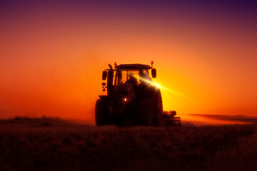 Farmer ploughs a field with tractor in sunset.  The tractor is backlit by the setting sun. 