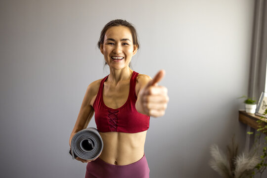 Happy Asian girl holding yoga mat and looking at camera u dnevnoj sobi. Smiling woman looking forward to fitness workout and getting ready to go to the gym. He points his thumb up