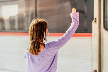 A young woman in a mask waves to somebody in the train from the platform of the train statinon