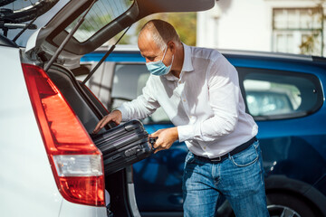 A taxi or Uber driver in mask puts the luggage into the trunk of the car