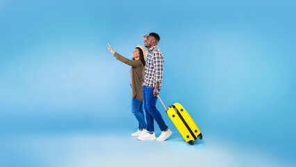 Cheery millennial black couple walking with suitcase, traveling together, having summer vacation on blue studio background