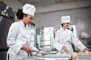 Two professional female chefs in white cook uniforms and aprons knead pastry dough and eggs, prepare bread, cookies, and fresh bakery food, baking in oven at a stainless steel kitchen of a restaurant.