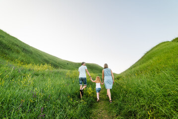 Fototapeta premium Mom, dad, and daughter walk back in the green grass. Happy young family spending time together, running on nature, on vacation, outdoors. The concept of a family holiday. Back view.
