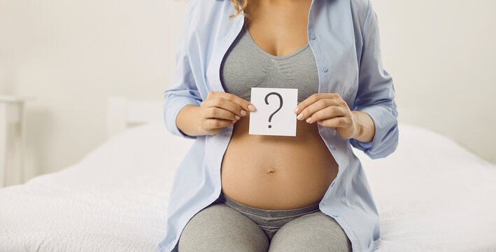 Pregnancy and thinking. Young pregnant woman holding small piece of paper with question mark guesses sex of child or chooses name. Cropped image of unknown woman with pregnant tummy sitting on bed.