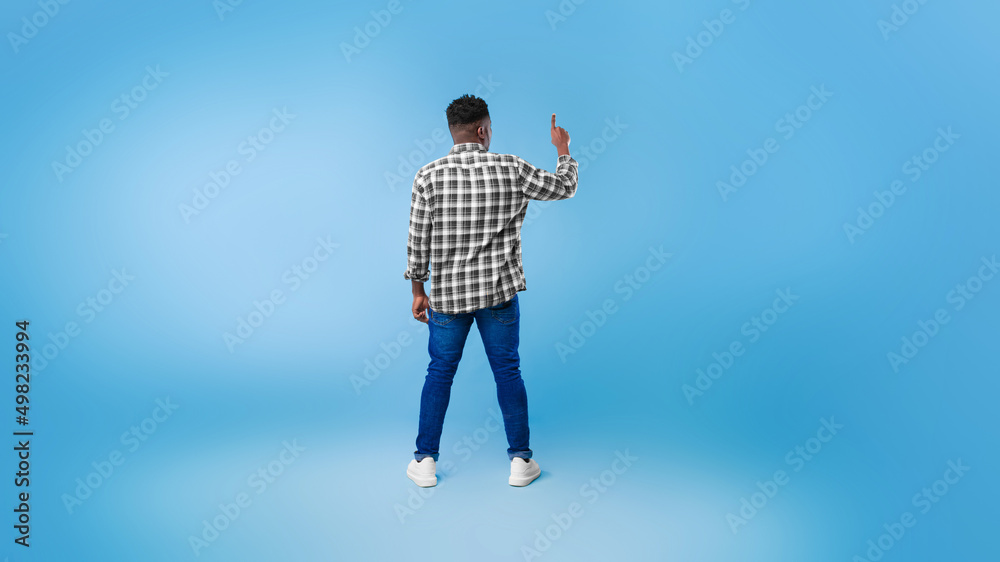 Poster back view of young black guy pushing button on virtual screen over blue studio background, mockup fo