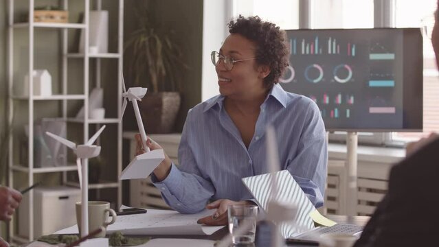 Waist up of multiethnic team of engineers sitting at office table discussing alternative energy sources. African American woman holding model of wind turbine while talking then handing it to others
