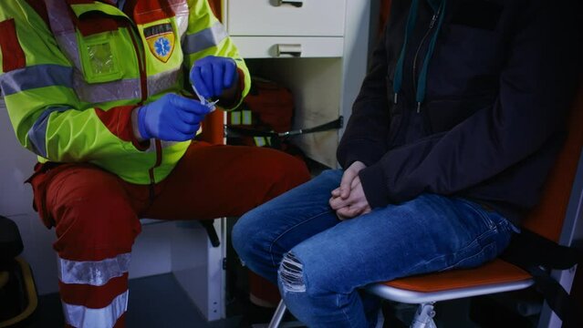Medical Emergency Care Worker Sitting At the Back Of an Ambulance Car Next to the Young Caucasian Guy, Taking Out a Q-Tip To Test The Person For COVID-19.