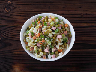 Traditional vegetable salad olivier with salted cucumbers in a white bowl on a dark wooden table, top view, flat layout. Mixed salad recipe for lunch, russian cuisine