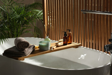 Wooden bath tray with candle, air freshener and bathroom amenities on tub indoors