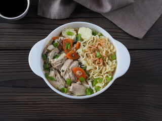 Chicken soup with chinese noodles, vegetables and meat in asian style in a white bowl, soy sauce, napkin on a dark wooden table, flat layout. Broth with instant vermicelli. Oriental cuisine