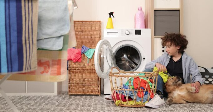 Housework. Boy doing laundry at home loading clothes into washing machin and playing with dog.