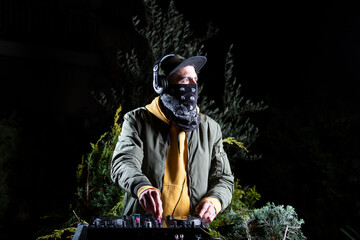 DJ chico play musica con una mesa de mezclas en una fiesta en bosque vestido con una chaqueta verde, una sudadera con capucha y una gorra, rave party © Alex