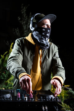 DJ vertical chico pincha musica con una mesa de mezclas en una fiesta en bosque vestido con una chaqueta verde, una sudadera con capucha y una gorra, rave party