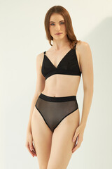 Obraz premium Fashion photo of young lady with perfect body in a black designer lingerie. Girl on a sunny day sunbathes in sexy pose sitting on the white background.