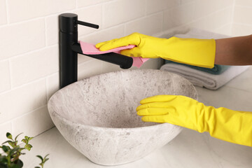 Woman in gloves cleaning faucet of bathroom sink with rag, closeup