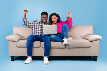 Young black couple sitting on sofa with laptop, raising hands up in excitement, celebrating success on blue studio background