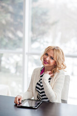Fototapeta premium Happy beautiful blonde woman with tablet sitting at table in co-working space, looking at camera and smiling. Medium shot. Wireless communication concept