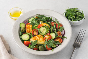 Halloumi salad with arugula, tomato, cucumber, onion and olives in bowl on light background. Mediterranean dish with fried cheese and vegetables