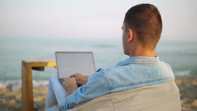 Young Successful Man Freelancer Typing And Coding On Laptop While Lying On A Lazy Bag On The Tropical Beach At Sunrise, Working Remotely During Summer Vacation. Distance And Pandemic Work Concept