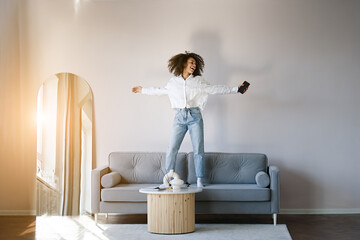 Carefree joyful african american girl jumping dancing alone moving to rhythm in living room, happy african American young woman have fun enjoying listening to music, entertain on weekend at home