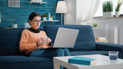 Happy student listening to online class lesson on laptop at home, sitting on couch and attending remote school conference meeting. Cheerful person looking at computer screen in living room.