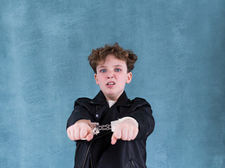 Medium horizontal shot of grimacing boy in black leather jacket handcuffed against turquoise...