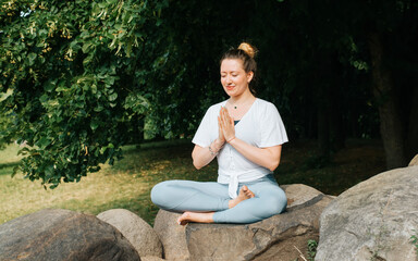 Meditation and yoga, healthy lifestyle concept. Smiling caucasian woman with closed eyes meditating in nature. Yogi woman sitting in lotus position on stones outdoors