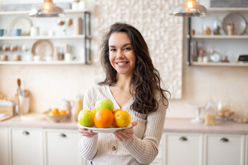 Happy woman holding plate with fresh fruits, looking and smiling at camera, standing in kitchen interior