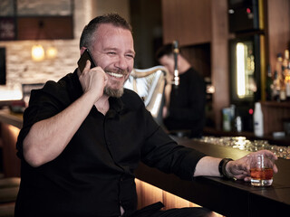 Portrait of an attractive happy man holding a glass of whiskey and talking on his mobile phone at the bar or pub.