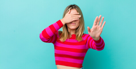 young pretty blonde woman covering face with hand and putting other hand up front to stop camera,...