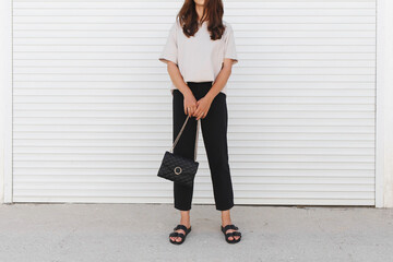 Woman wearing beige t-shirt, black pants, bag with chain and flat sandals walking outdoor near white roller door. Details of stylish trendy basic minimalistic casual outfit. Street fashion. No face.