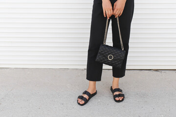 Bag in women's hands. Woman wearing black pants, bag with chain and flat sandals standing outdoor. Details of stylish trendy basic minimalistic casual outfit. Street fashion. Women's legs, no face.