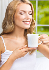 Portrait  image of happy smiling woman with cup near window. Amazed blond girl with closed eyes drinking coffee indoors. Morning calm concept.