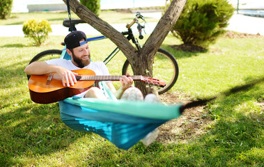 bearded man with a guitar lying in a hammock smiling against the backdrop of nature and greenery.