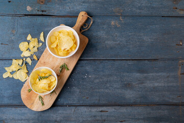 Overhead view of potato chips in bowl with rosemary on serving board at wooden table