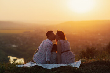 Husband kisses his wife while they sitting on the hill at sunset. Little daughter sits between the...