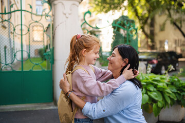 Happy schoolgirl hugging his grandmother waiting for her after school outdoors in street.