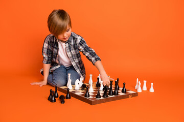 Full body photo of young boy sit floor play game clever learn chess isolated over orange color background