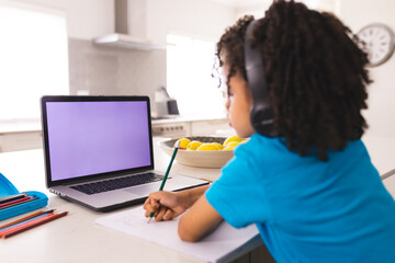 Hispanic boy wearing headphones while writing and attending online class through laptop