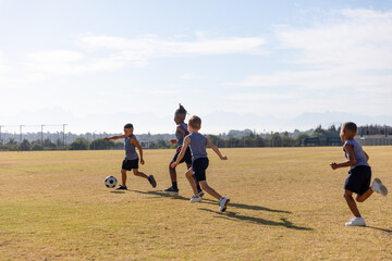 Multiracial elementary schoolboys playing soccer on school soccer field against sky during sunny day